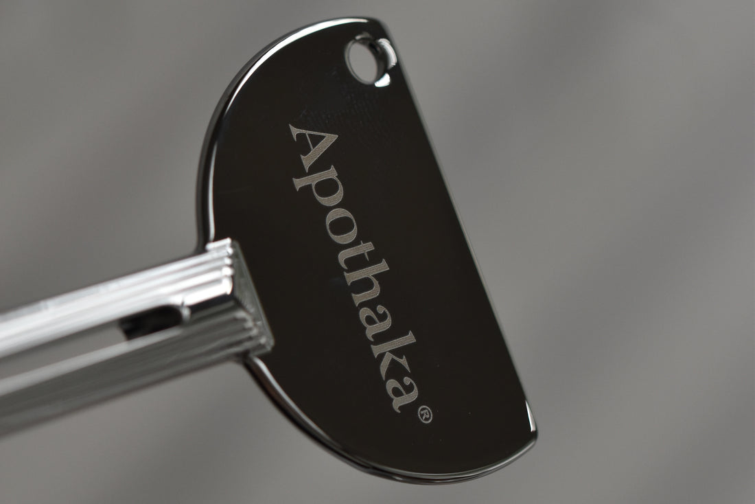 A close-up of a metal tube squeezer key with the brand name 'Apothaka' printed vertically on a polished dark metal face, set against a soft gray background.