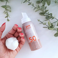 A hand with pink nail polish holds a golf ball-size amount of EVY daily defence SPF50 face mousse , styled with green eucalyptus branches on a white background.